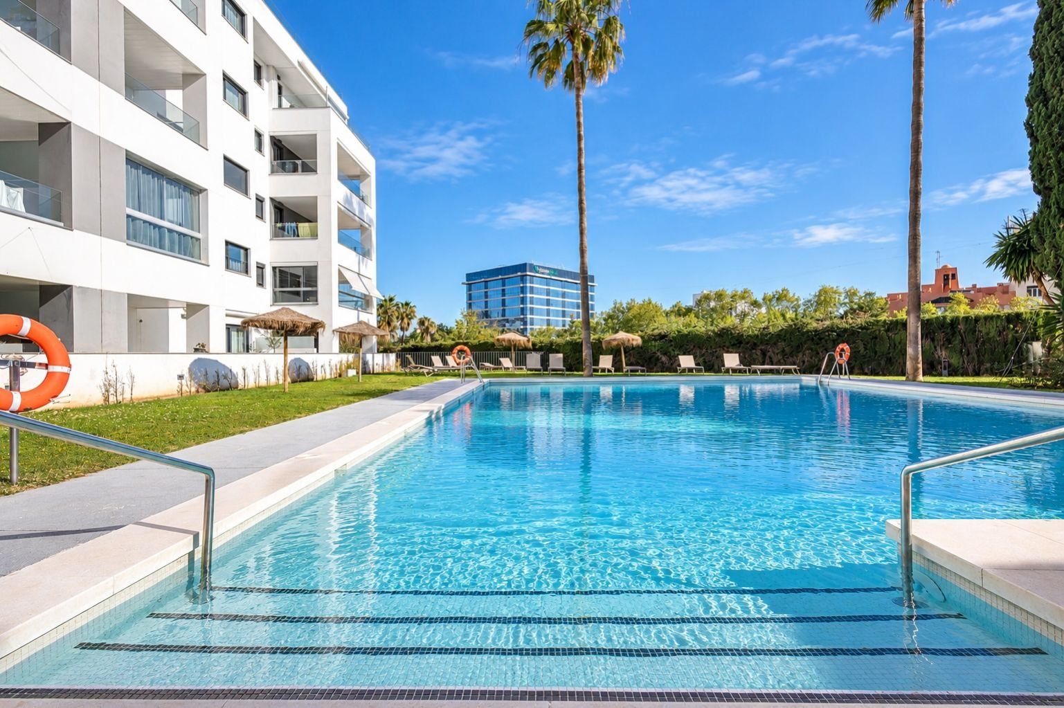 A swimming pool with clear blue water, surrounded by a white building with balconies, palm trees, and a clear sky.