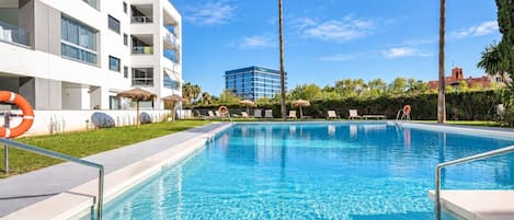 Une piscine à l’eau d’un bleu cristallin, entourée d’un bâtiment blanc doté de balcons, de palmiers et surplombant un ciel dégagé.