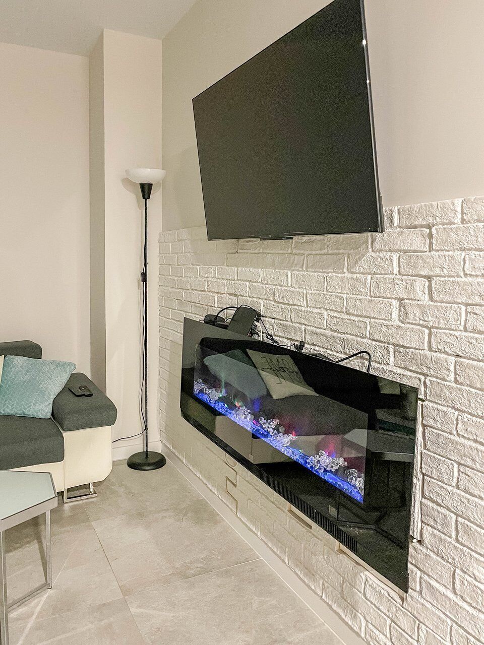 A modern living room with a flat-screen TV mounted above a white brick fireplace, a sofa with cushions, and a floor lamp.