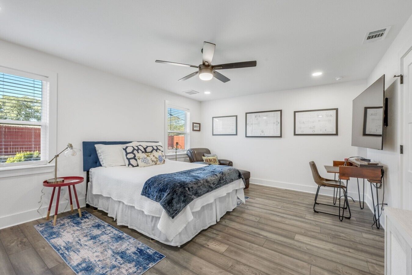 A bedroom with a bed, a desk, a chair, a ceiling fan, and framed pictures on the wall.