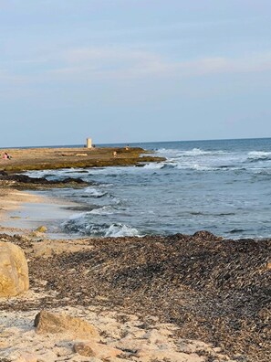 A rocky beach with waves, a lighthouse, and a clear sky.