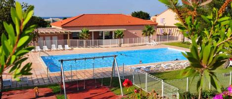 Una piscina con un edificio de techo rojo al fondo, rodeada de sillas reclinables blancas y una zona vallada con flores.