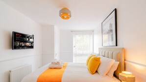 A hotel room with a bed, a bedside table with a lamp, a wall-mounted TV displaying Netflix content, a window with blinds, and a framed picture on the wall. - 2-Bed Flat Sleeps 4 with Parking · Walk to Historic Dockyard Chatham (Gillingham)