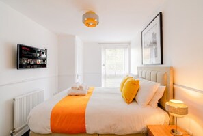 A hotel room with a bed, a bedside table with a lamp, a wall-mounted TV displaying Netflix content, a window with blinds, and a framed picture on the wall. - 2-Bed Flat Sleeps 4 with Parking · Walk to Historic Dockyard Chatham (Gillingham)