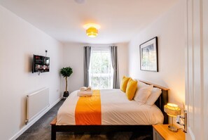 A hotel room with a bed, bedside lamp, window with curtains, radiator, and a wall-mounted television. - 2-Bed Flat Sleeps 4 with Parking · Walk to Historic Dockyard Chatham (Gillingham)