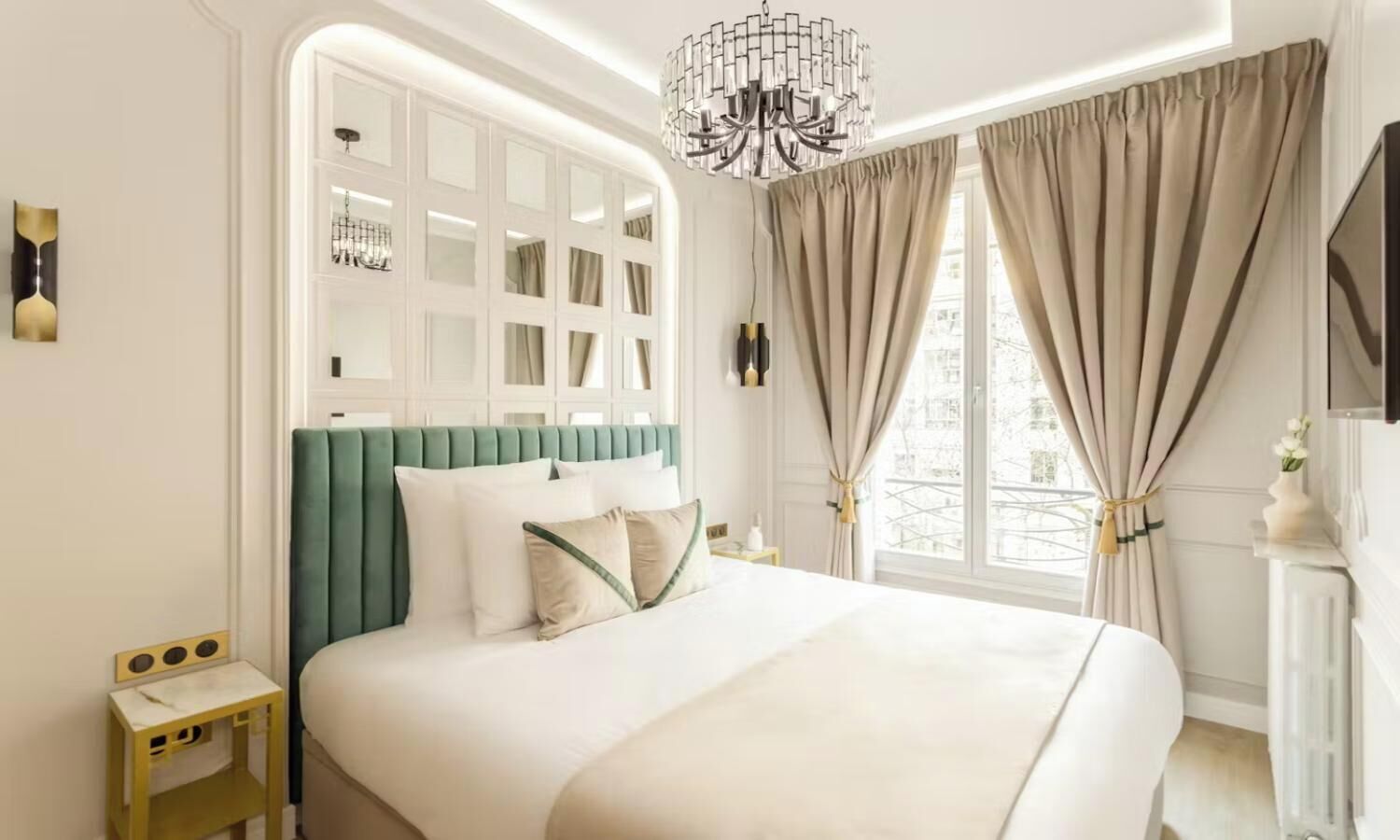 Ein Hotelzimmer mit einem großen Bett, einem mit grünem Stoff bezogenen Kopfteil, einer Kristallleuchtre und Blick auf die Stadt durch das Fenster.
