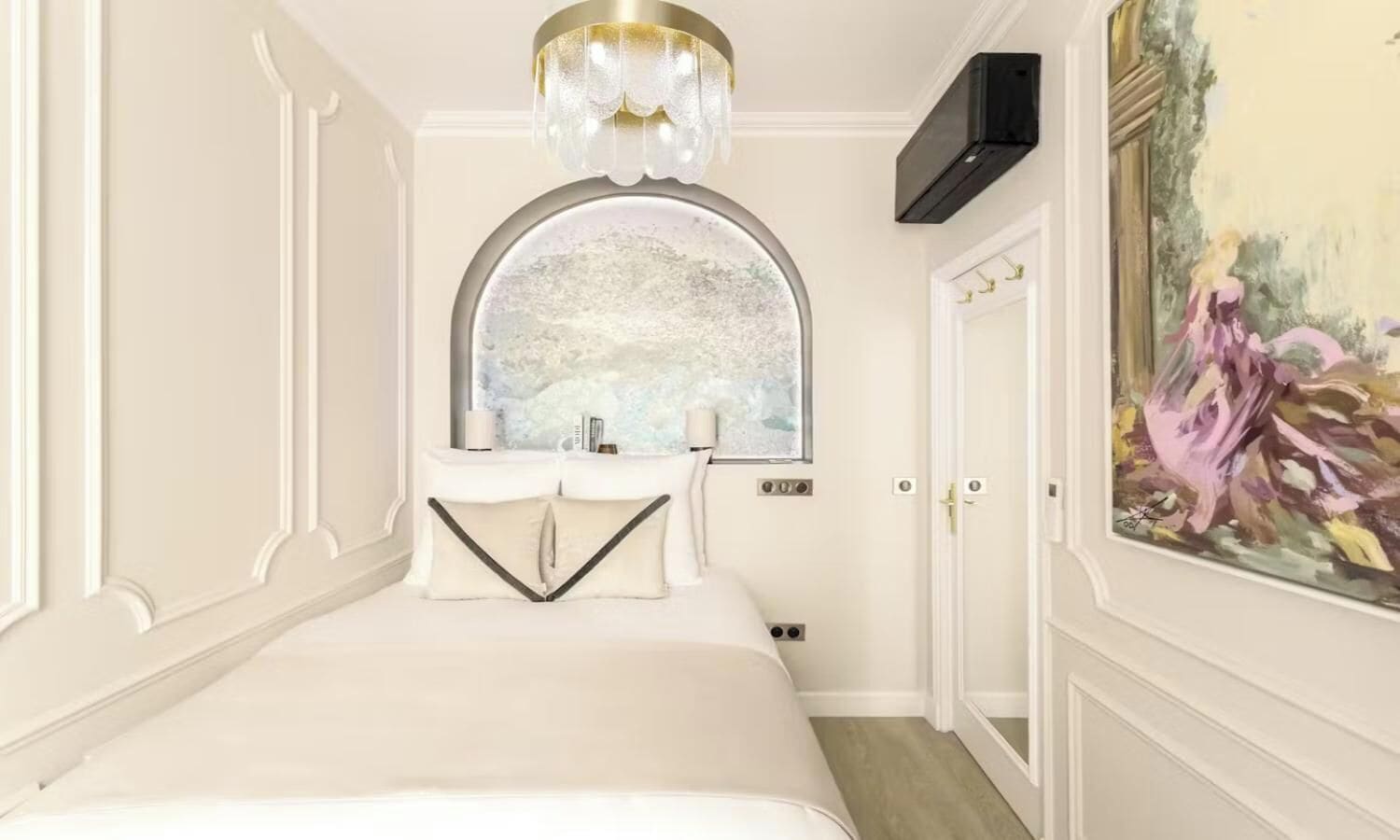 A hotel room with a large bed, a chandelier, a window with a view, and a painting on the wall.