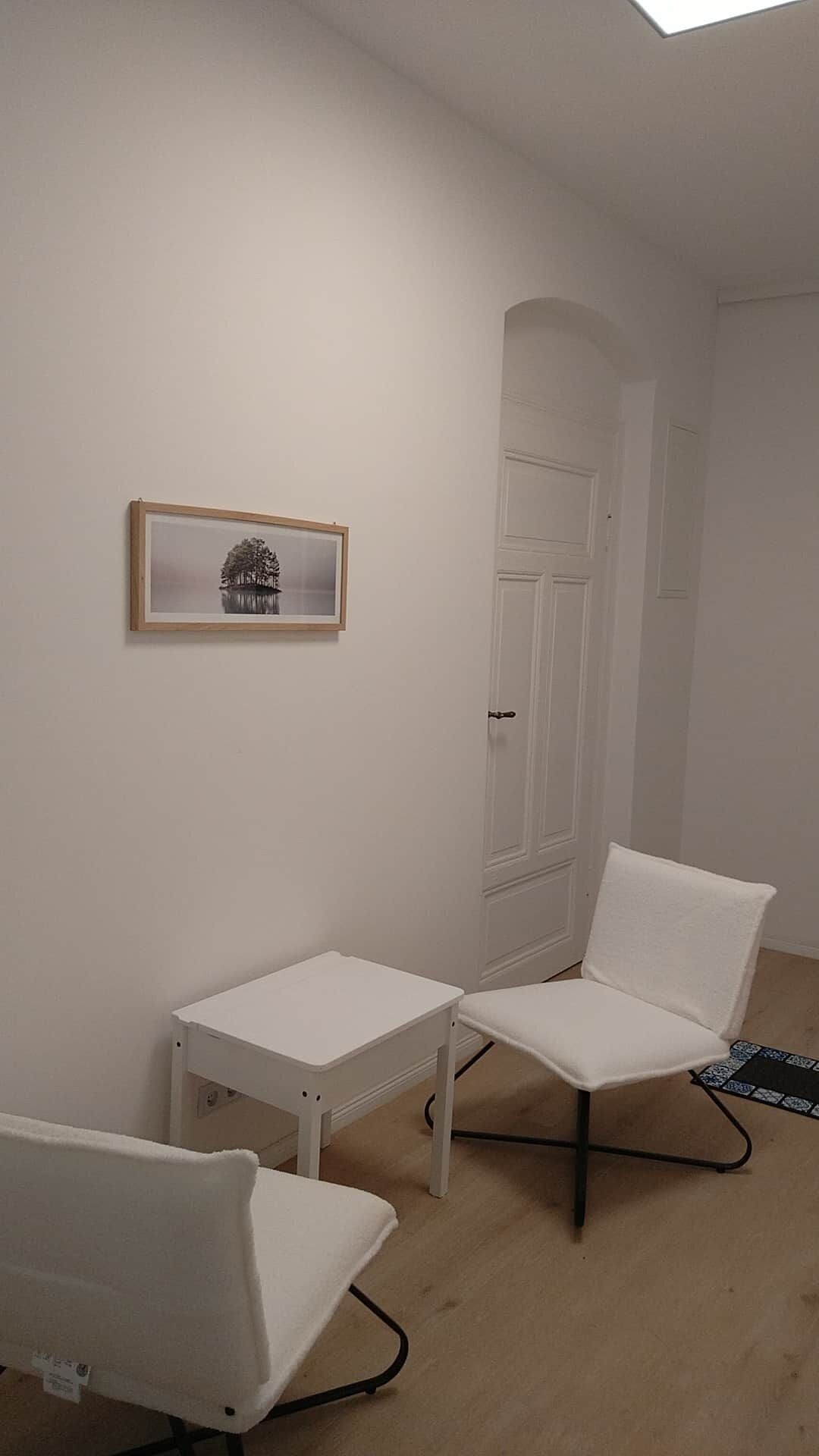 Ein Zimmer mit einer weißen Tür, einem gerahmten Bild an der Wand und zwei weißen Stühlen mit einem kleinen Tisch.