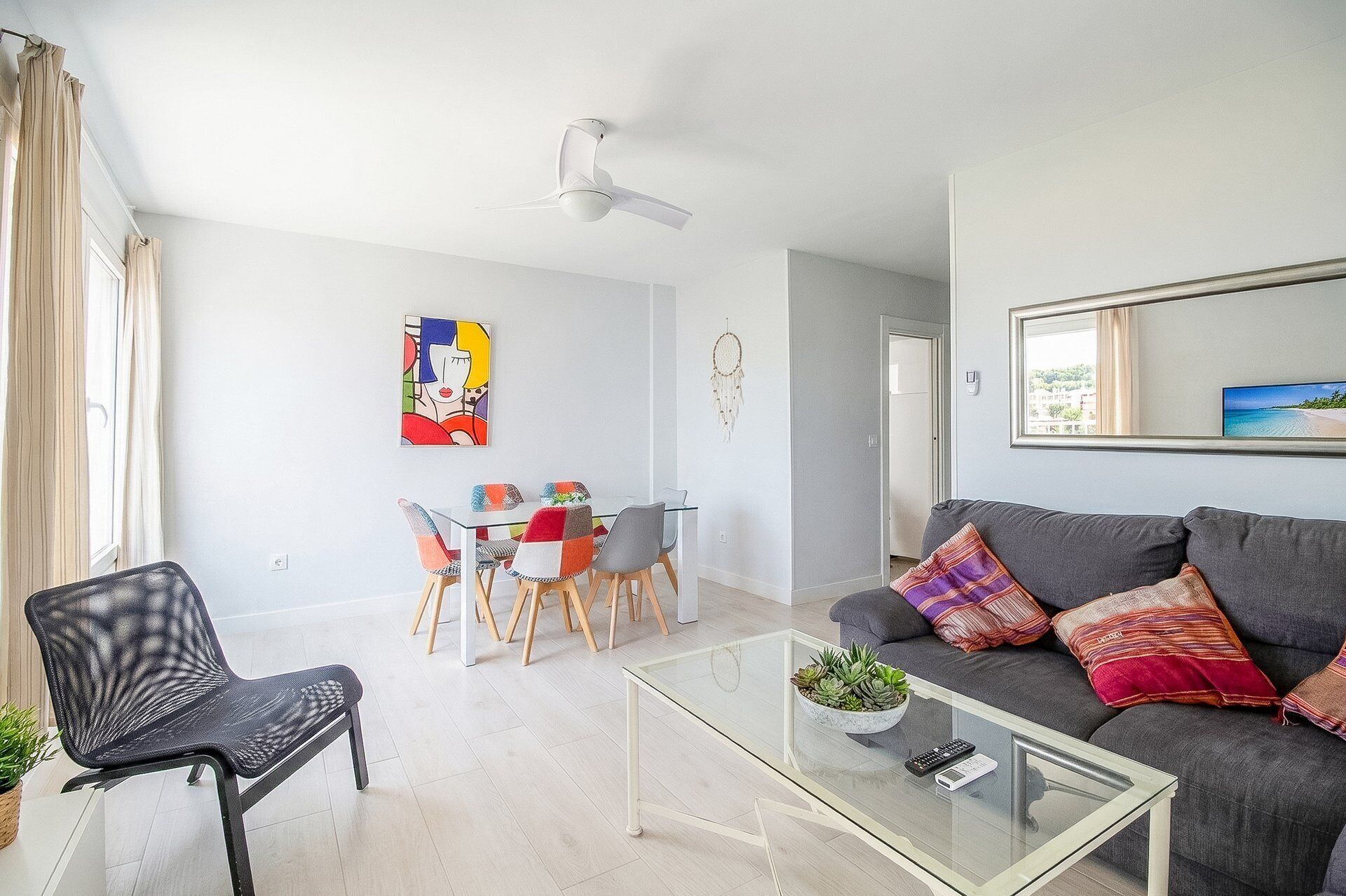 Uma sala de estar moderna com uma mesa de centro de vidro, sofá cinza com almofadas estampadas, área de jantar com mesa e cadeiras de vidro, ventilador de teto e obras de arte nas paredes.
