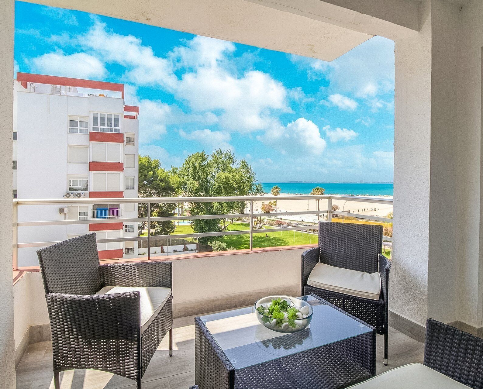 A balcony with wicker chairs and a glass table, overlooking a beach and a building with red accents.
