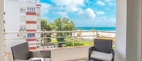 A balcony with wicker chairs and a glass table, overlooking a beach and a building with red accents.