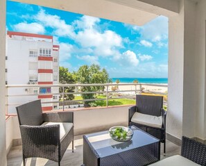 Ein Balkon mit Korbstühlen und einem Glastisch, mit Blick auf einen Strand und ein Gebäude mit roten Akzenten.
