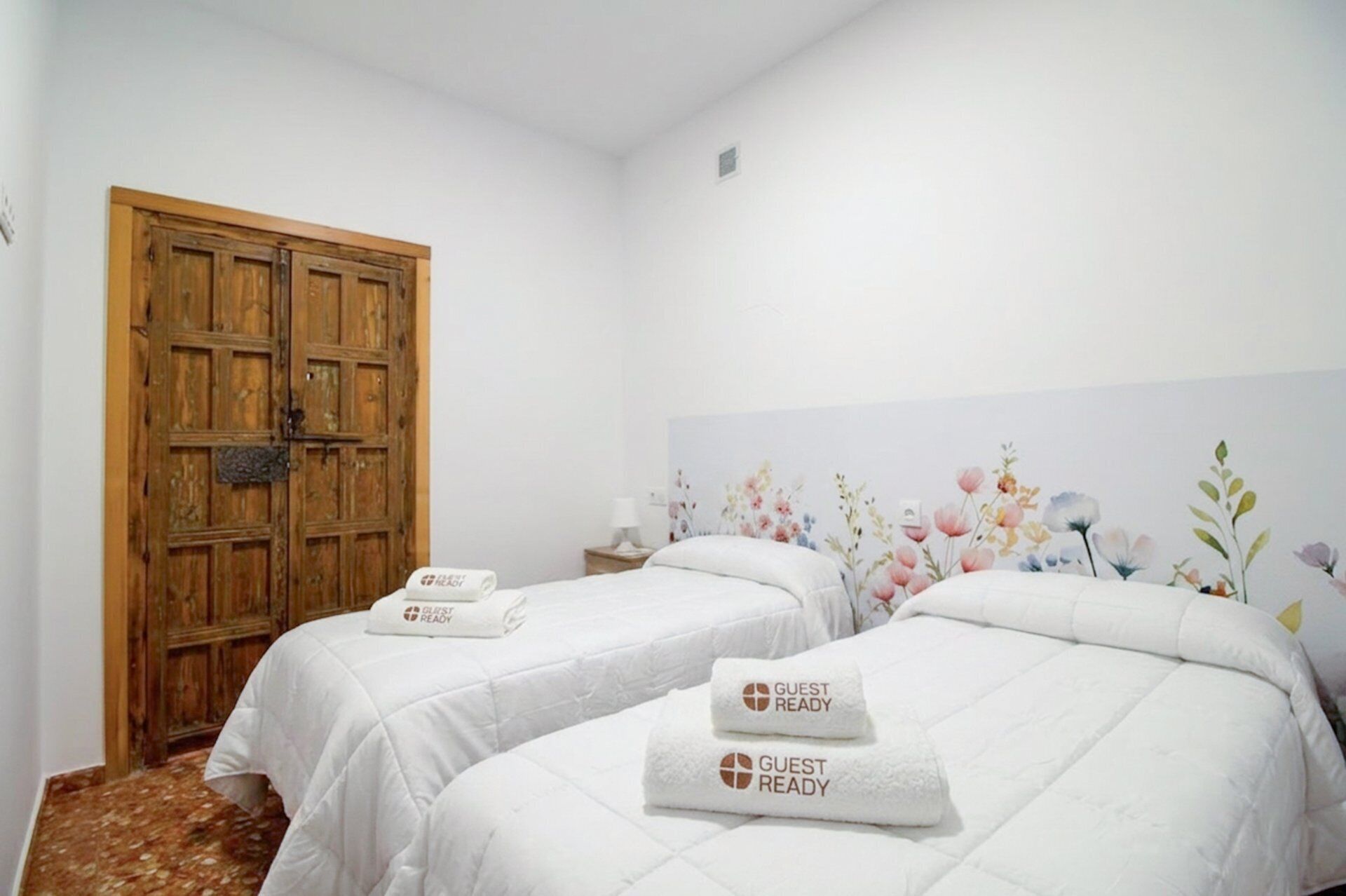 A hotel room with two beds, each with a white bedspread and a 'GUEST READY' towel. There's a wooden door and a floral wall decoration.