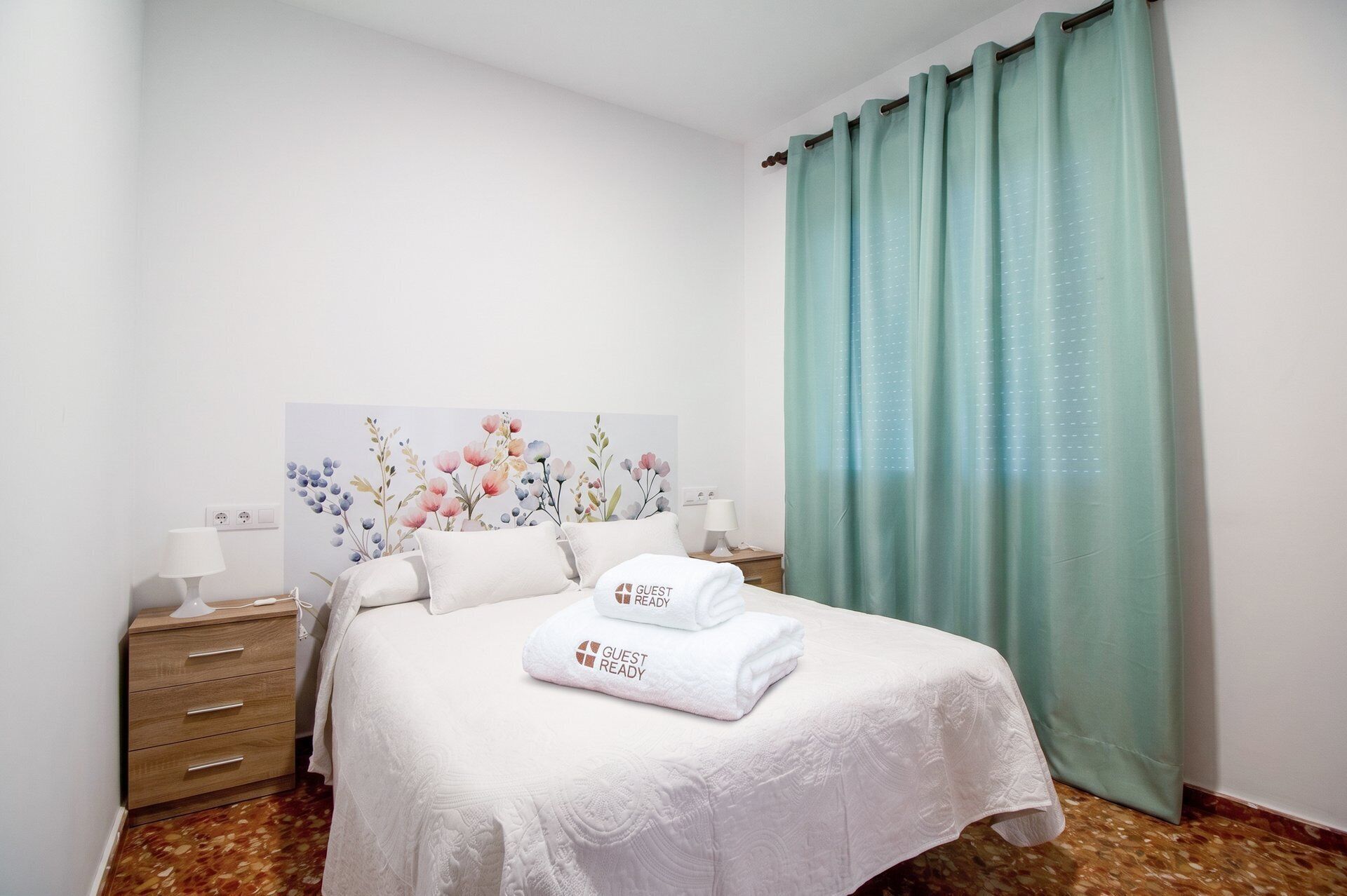 A hotel room with a bed, two bedside tables, a wall-mounted lamp, a painting, and a green curtain.