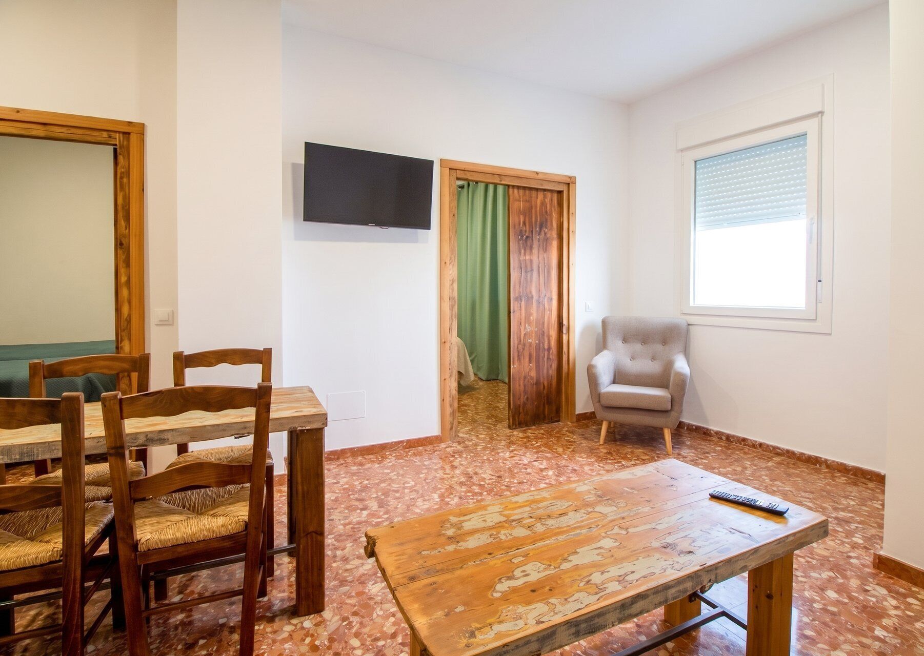 A room with a wooden dining table, chairs, a television mounted on the wall, a window with blinds, and a door with a green curtain.