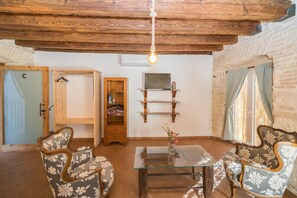 Ein gemütliches Wohnzimmer mit Holzboden, einem Glastisch, blumengemusterten Stühlen, einem Holzregal und einem Fernseher auf einem Holzbodenständer.