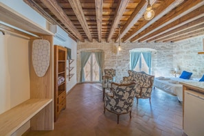Ein rustikales Zimmer mit freiliegenden Holzbalken, einer Steinwand, einem Holzschrank, einem blumigen Sessel, einem kleinen Tisch mit Lampe und einem Bett mit blauen Kissen.