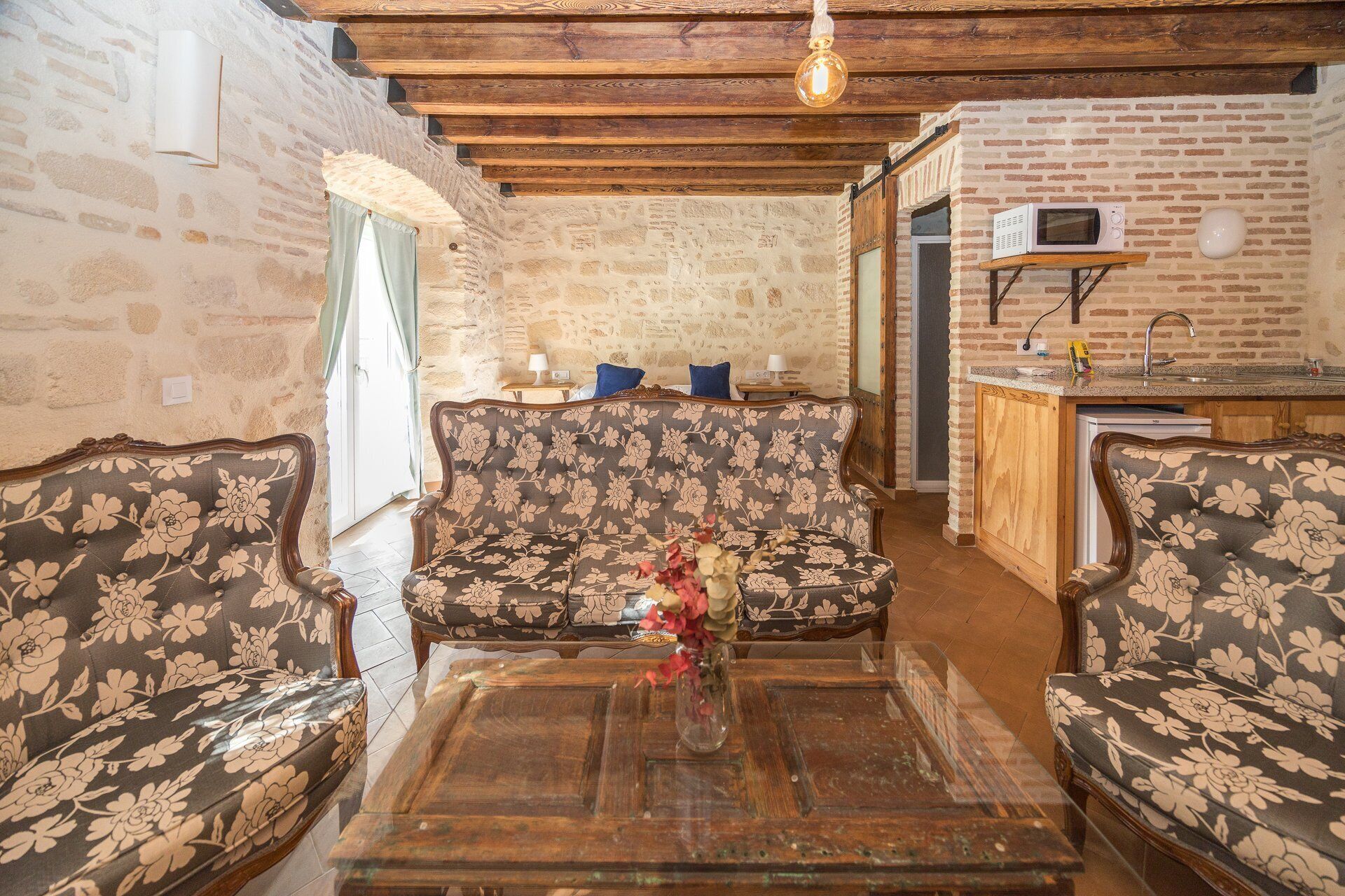 Ein rustikaler Innenraum mit einer Steinwand, Holzbalken, einer floralen Sofagarnitur, einem Holztisch und einer Küchenzeile mit einer Mikrowelle.