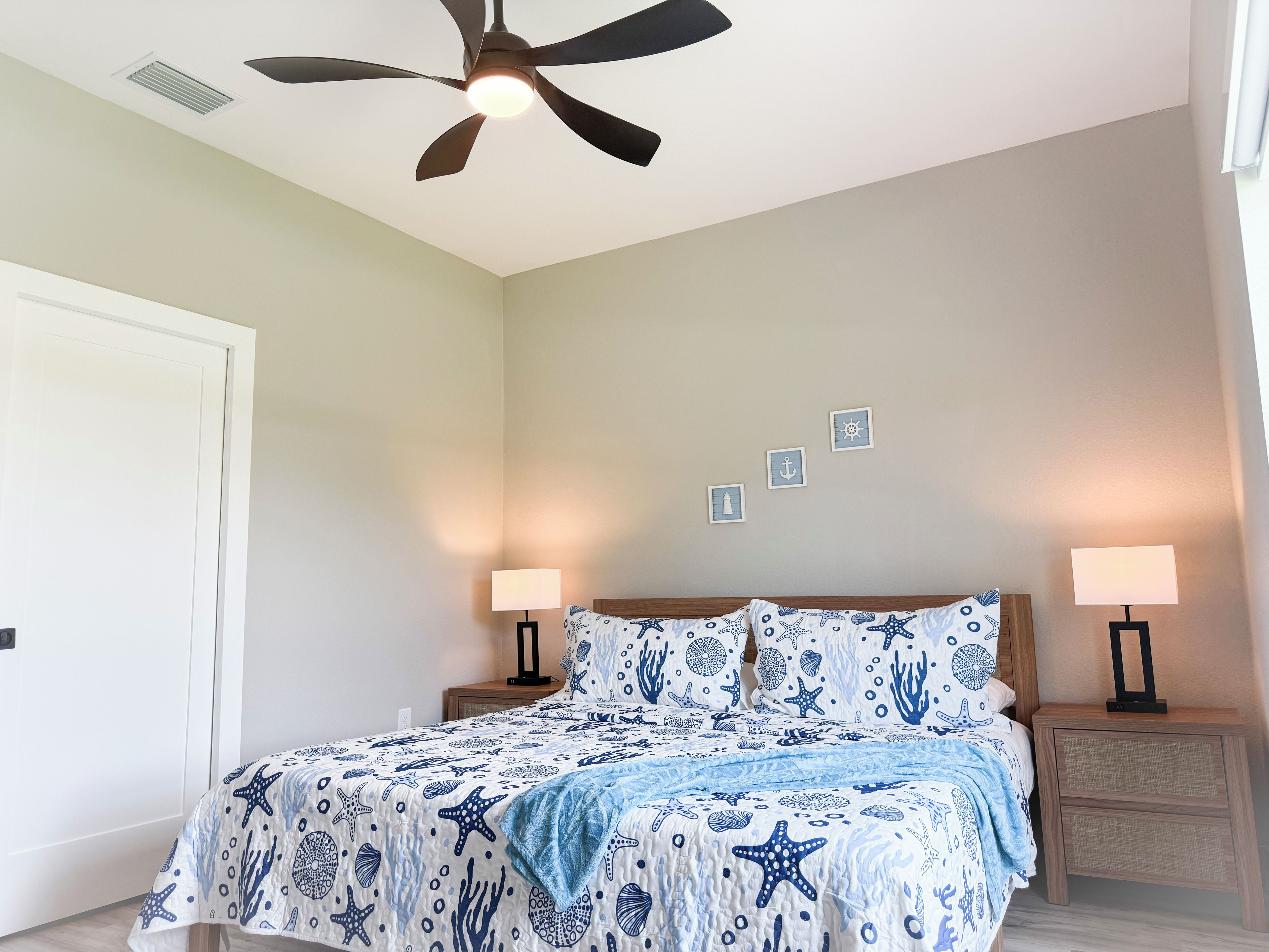 Quarto com cama, duas mesinhas de cabeceira, ventoinha no teto e obra de arte na parede.