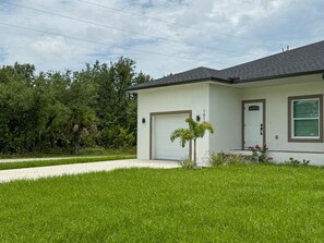 Una casa de un piso con fachada blanca, puerta de garaje y un pequeño jardín frontal con césped y una palmera.