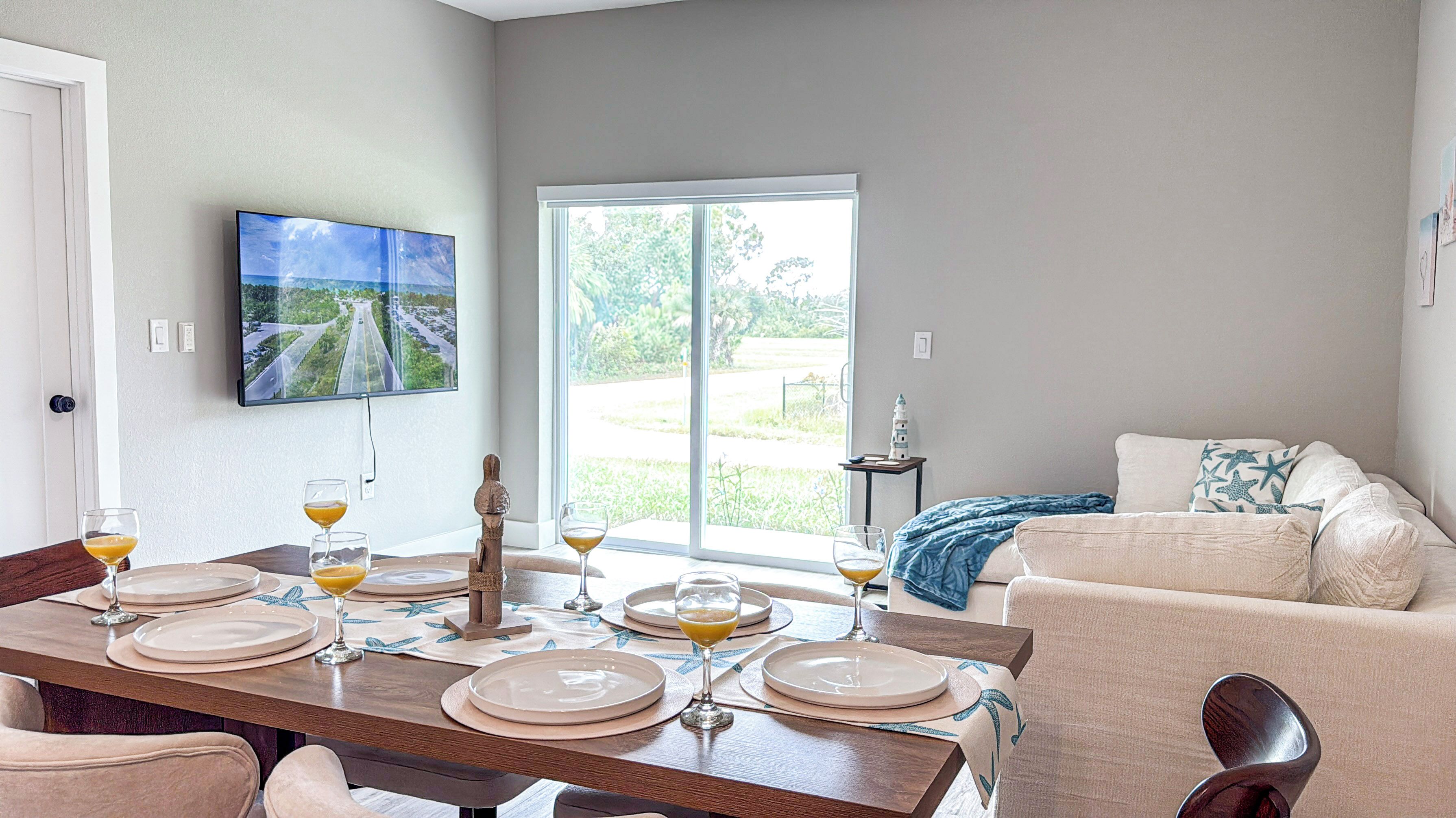 Un comedor con una mesa dispuesta para cuatro, un televisor montado en la pared y vistas a un paisaje verde a través de un amplio ventanal.