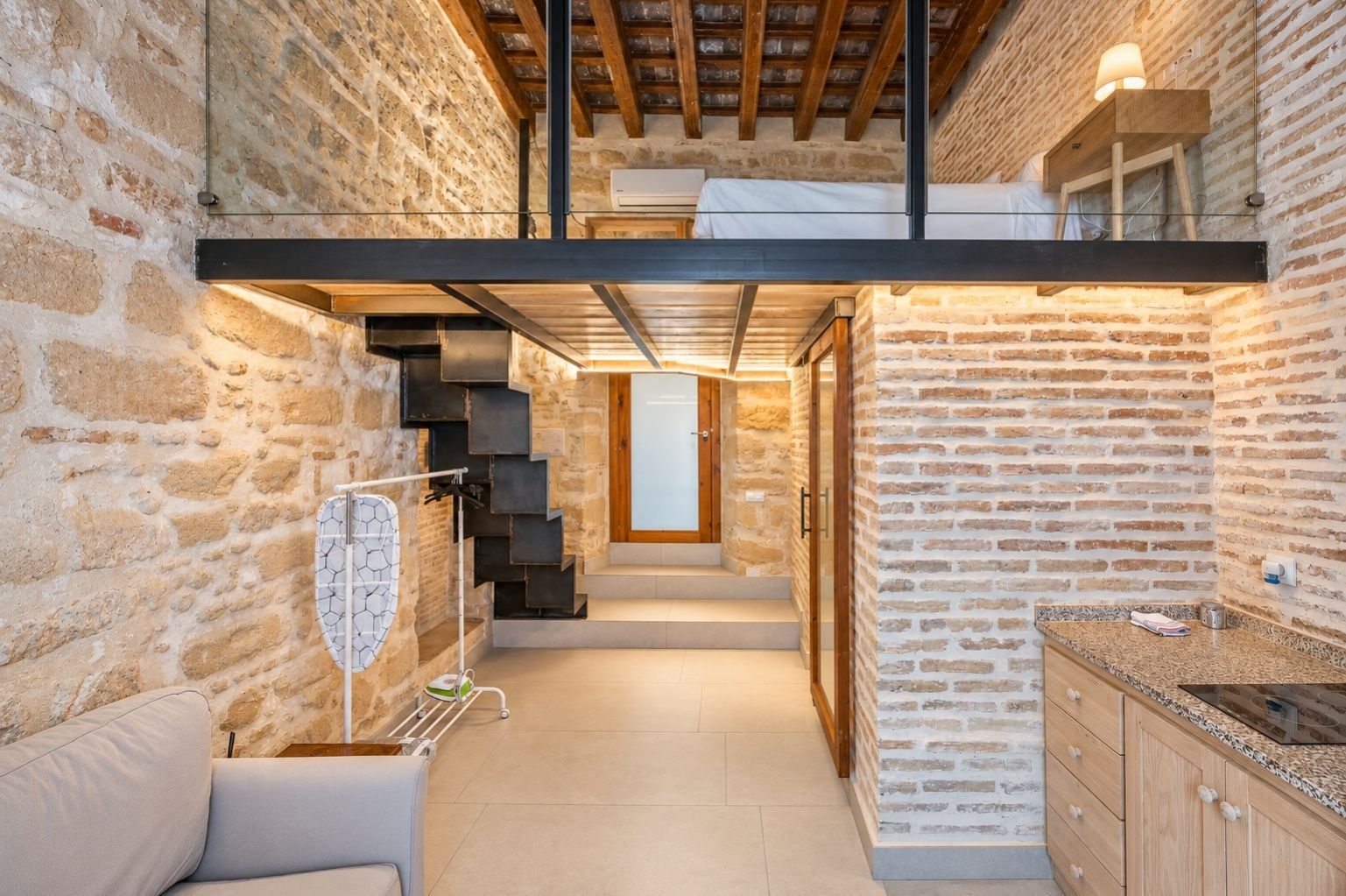 A modern interior with exposed brick walls, a staircase leading to a loft, and a kitchen area with a countertop and cabinets.