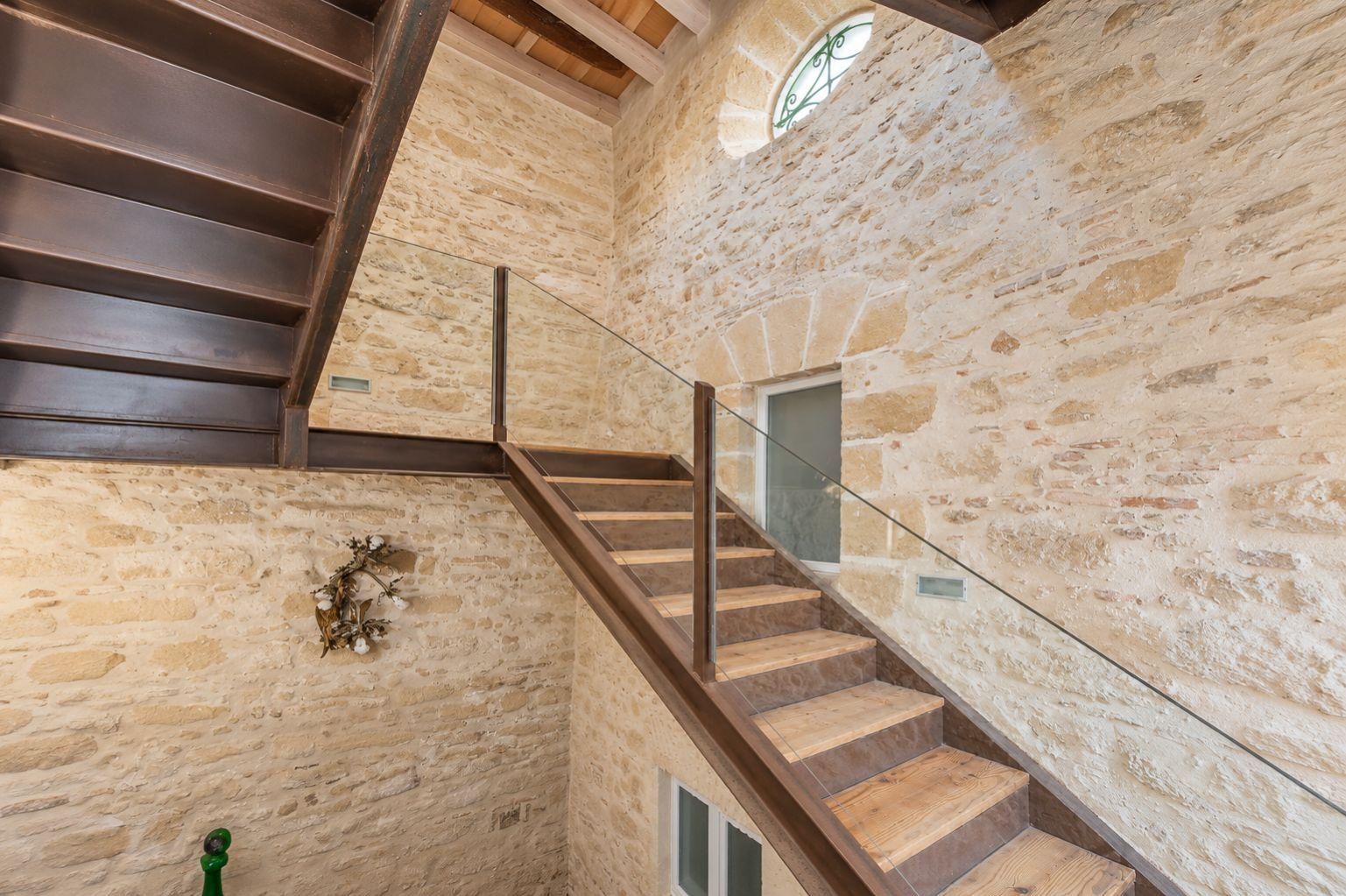 A staircase with wooden steps and glass railings, leading to a stone wall with a decorative wreath.