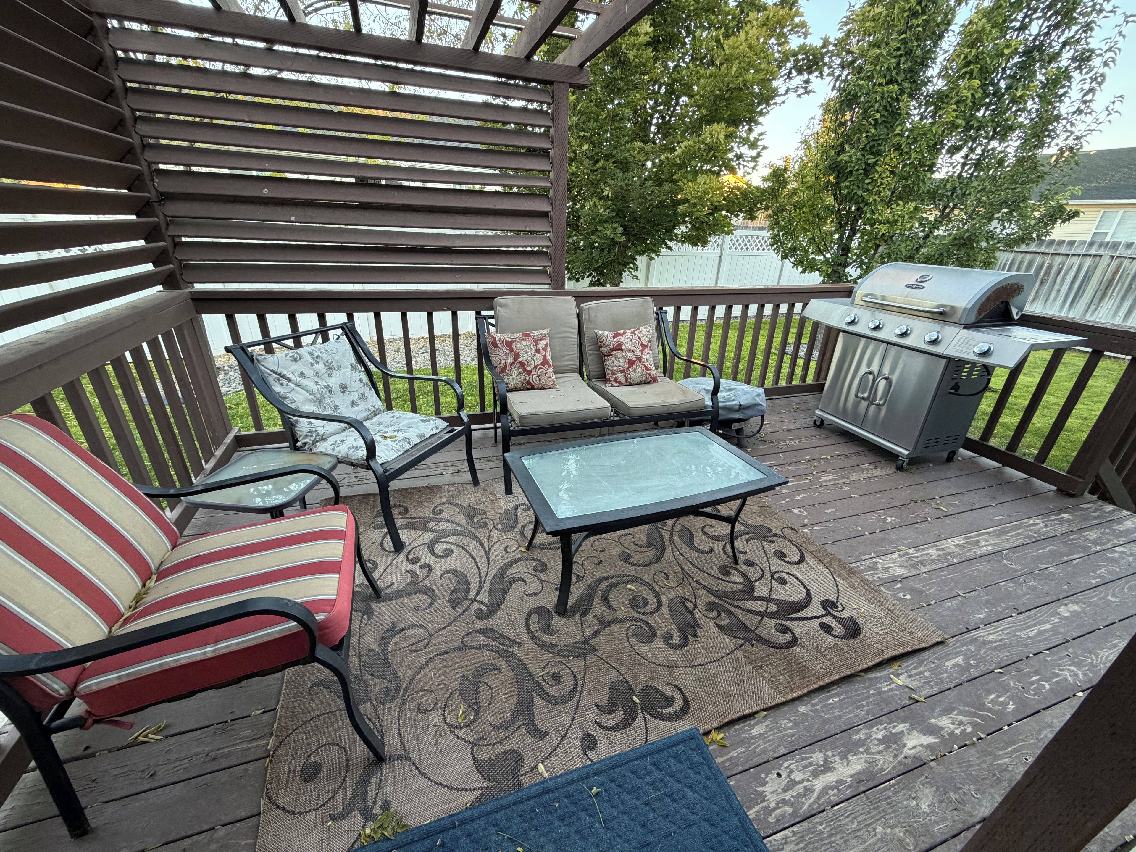 A covered outdoor patio with a grill, two cushioned chairs, a small table, and a patterned rug.