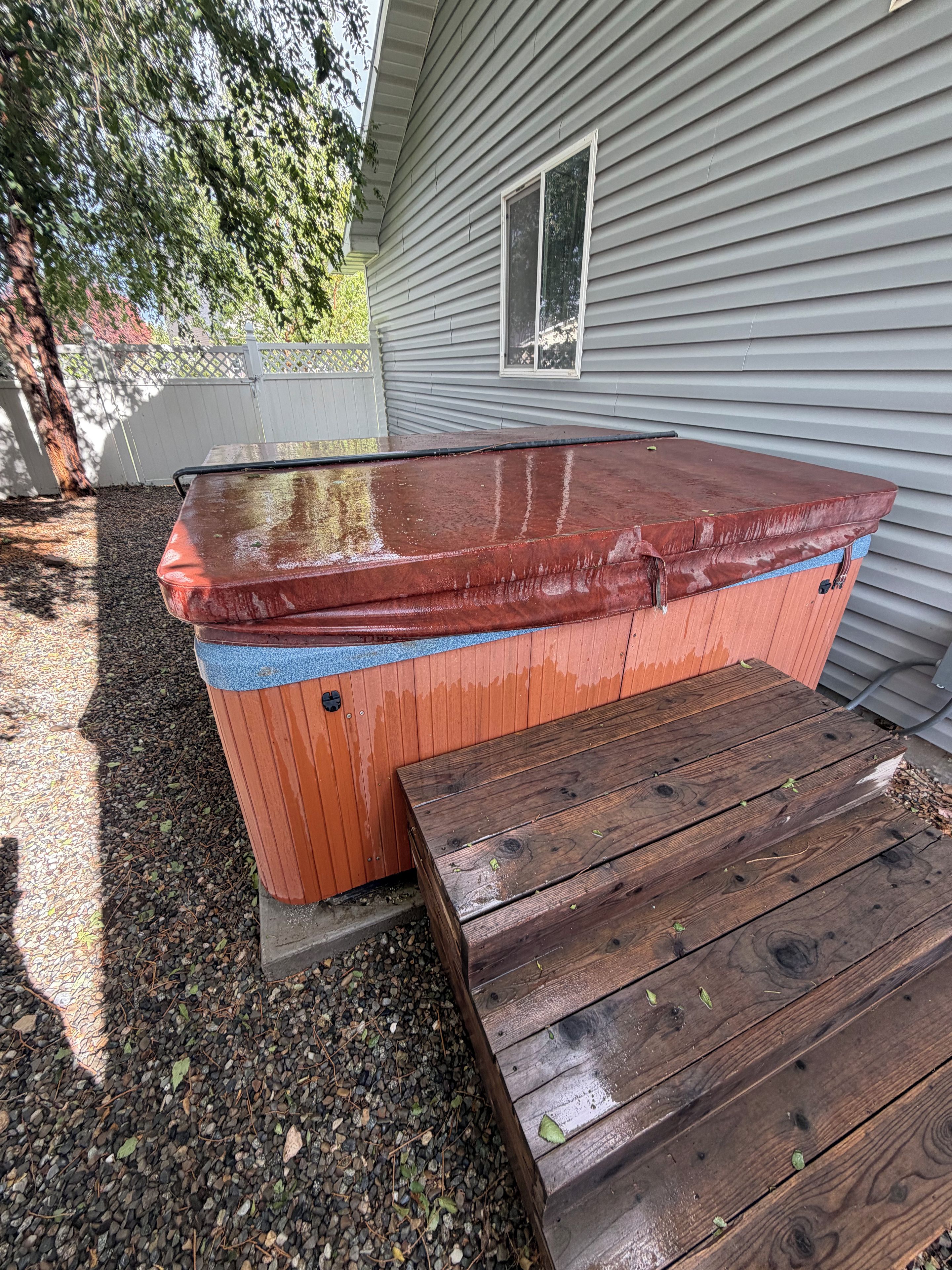 A hot tub with a wooden deck and steps, adjacent to a house with siding and a window.
