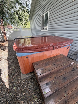 A hot tub with a wooden deck and steps, adjacent to a house with siding and a window. - Adventure Hub (Filer)