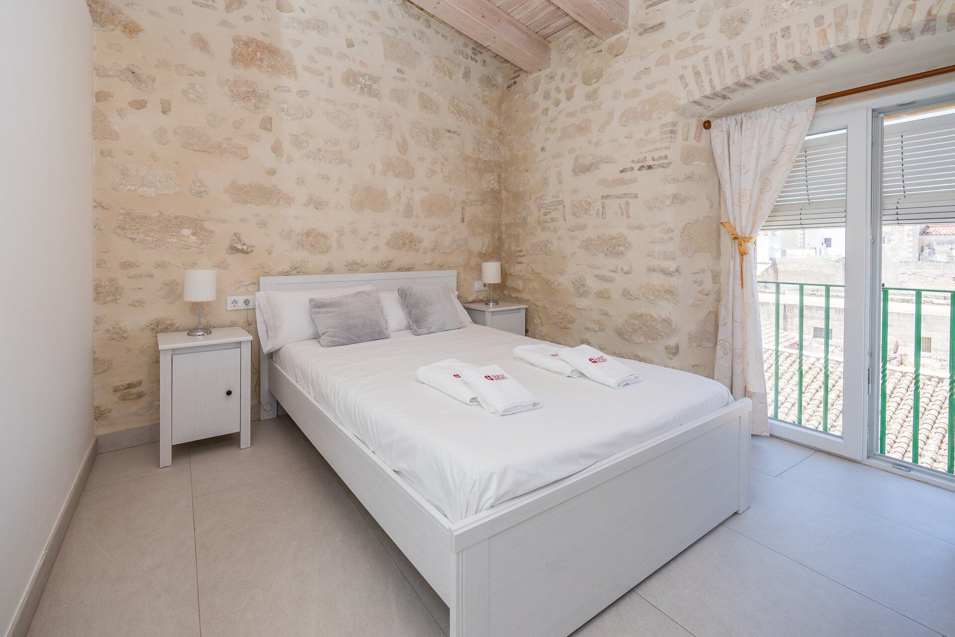 Una camera da letto con parete in pietra, un letto bianco, due comodini con lampade e un balcone con ringhiera verde.