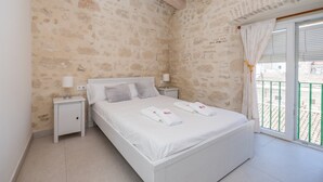 Un dormitorio con pared de piedra, una cama blanca, dos mesitas de noche con lámparas y un balcón con barandilla verde.