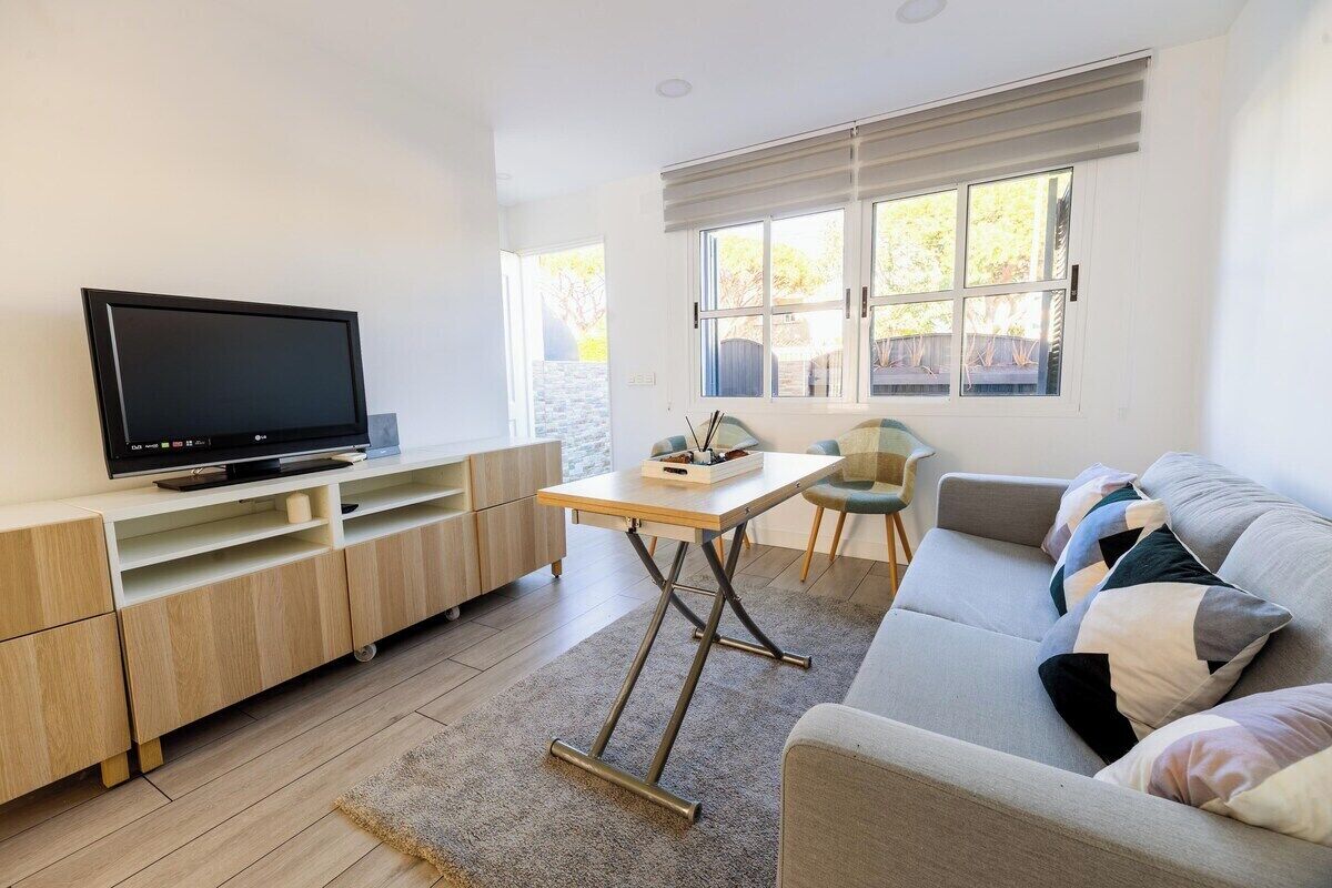 A modern living room with a flat-screen TV, a wooden entertainment unit, a grey sofa with patterned cushions, and a window with blinds.