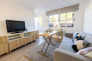 Ein modernes Wohnzimmer mit Flachbildfernseher, einer Holz-Unterhaltungsecke, einem grauen Sofa mit gemusterten Kissen und einem Fenster mit Jalousien.