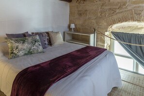 A bed with a white and purple bedspread, multiple pillows, a nightstand with a lamp, and a stone wall. - GuestReady - La Cruz Bajo A (El Puerto de Santa María)