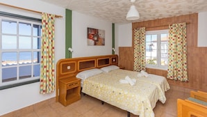 Ein Hotelzimmer mit Bett, Nachttisch, einem Fenster mit Vorhängen, einem Bild an der Wand und Blick auf das Meer.