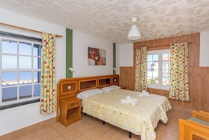 Ein Hotelzimmer mit Bett, Nachttisch, einem Fenster mit Vorhängen, einem Bild an der Wand und Blick auf das Meer.