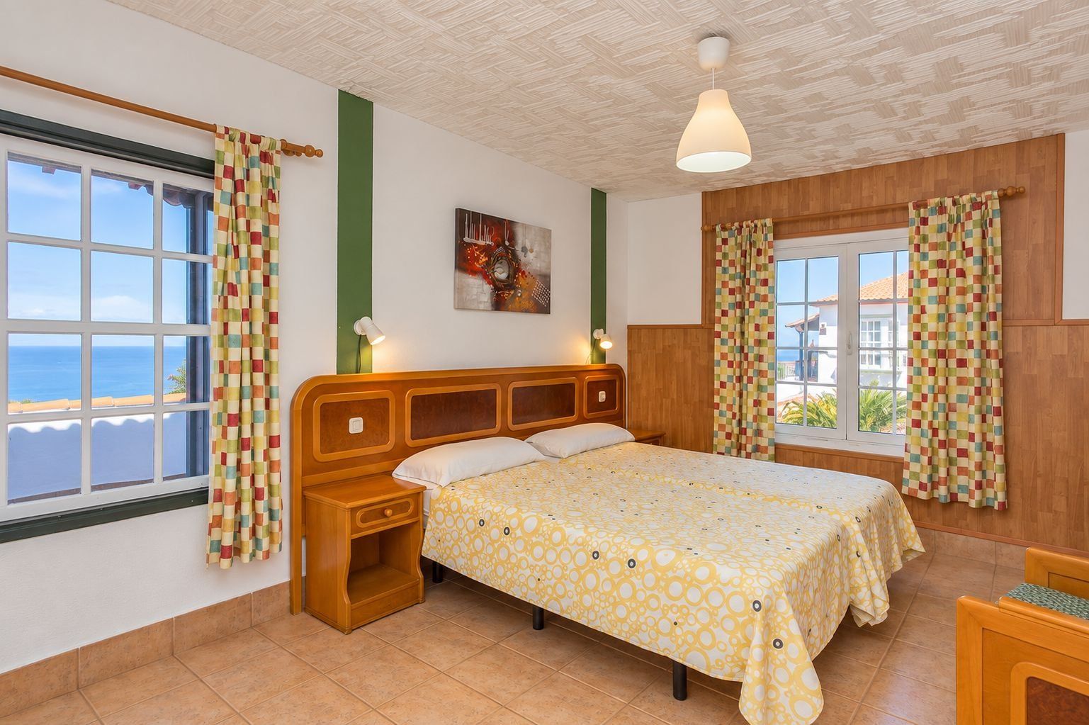 A bedroom with a large bed, a nightstand, a window with curtains, a painting on the wall, and a view of the sea.