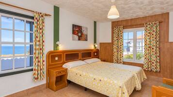 A bedroom with a large bed, a nightstand, a window with curtains, a painting on the wall, and a view of the sea.