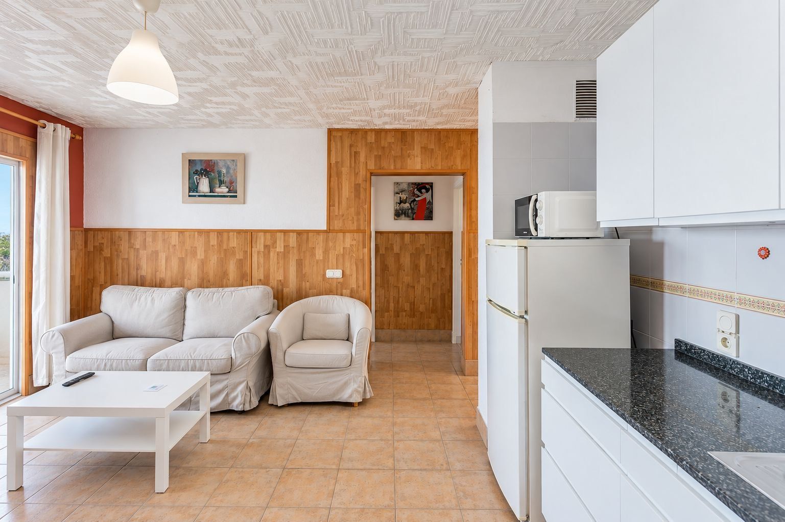 Un espacio habitable compacto con sofá, sillón y una mesita. Se observa una kitchenette con microondas y refrigerador.