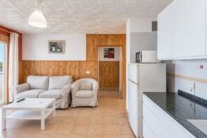 Ein gemütlicher Wohnbereich mit Sofa, Sesseln und einem kleinen Tisch. Eine Küchenzeile mit Mikrowelle und Kühlschrank ist sichtbar.