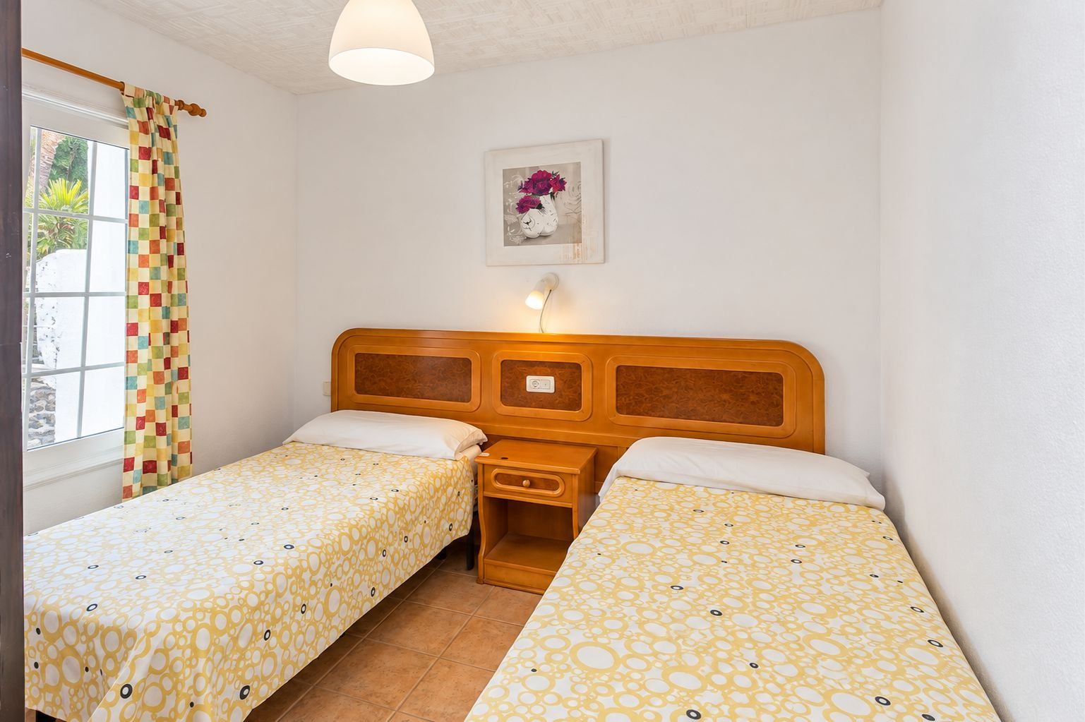 Una habitación con dos camas individuales, cabecera de madera, mesita de noche, ventana con cortina e imagen colgada en la pared.