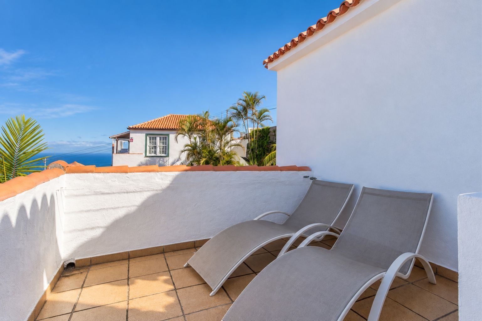 A balcony with two lounge chairs, a white wall, and a view of a house with a red roof.