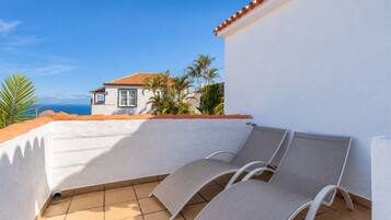 A balcony with two lounge chairs, a white wall, and a view of a house with a red roof.