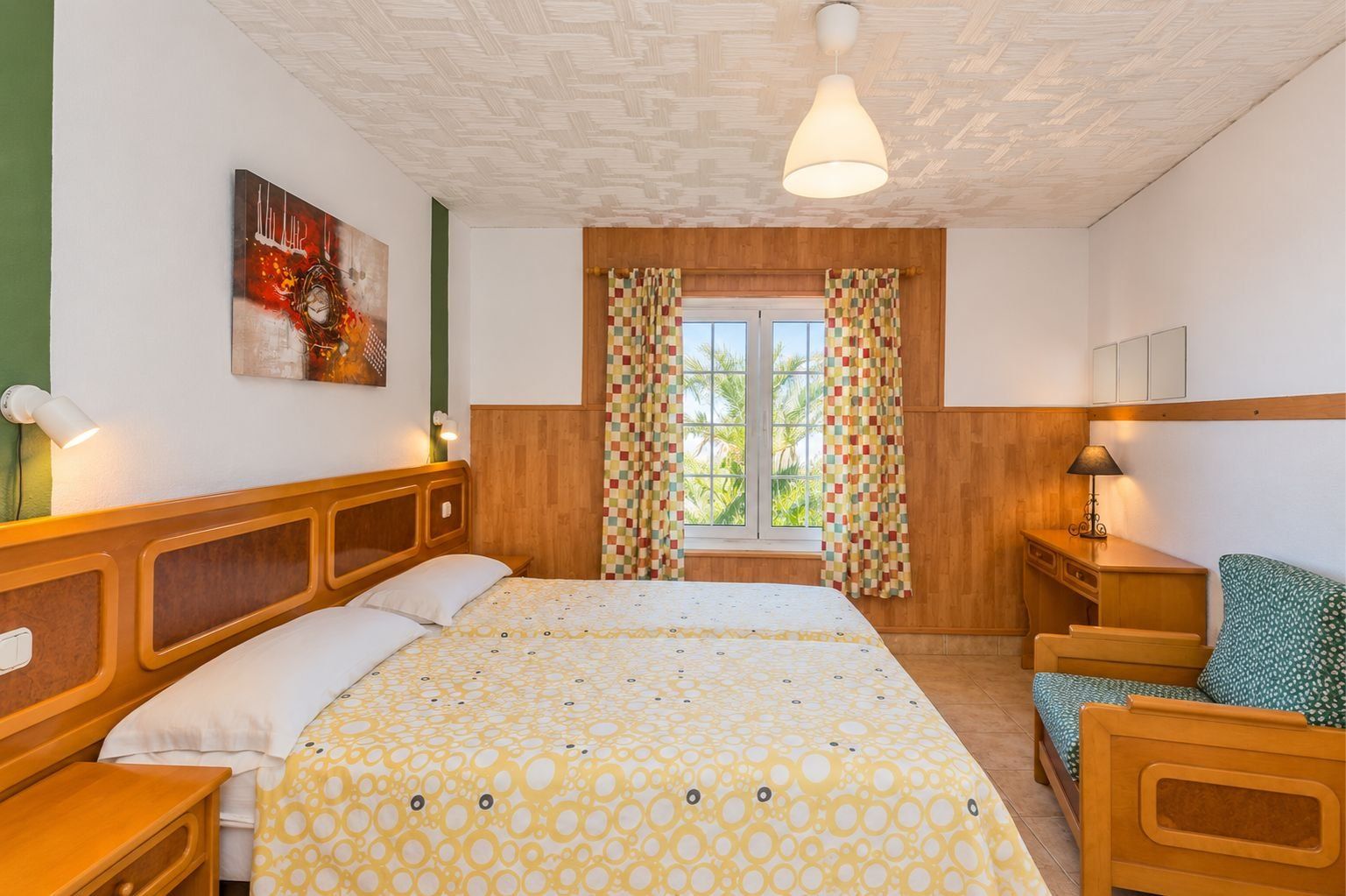 Ein Hotelzimmer mit Bett, Sessel, Schreibtisch, Lampe, einem Fenster mit Vorhängen und einem Bild an der Wand.