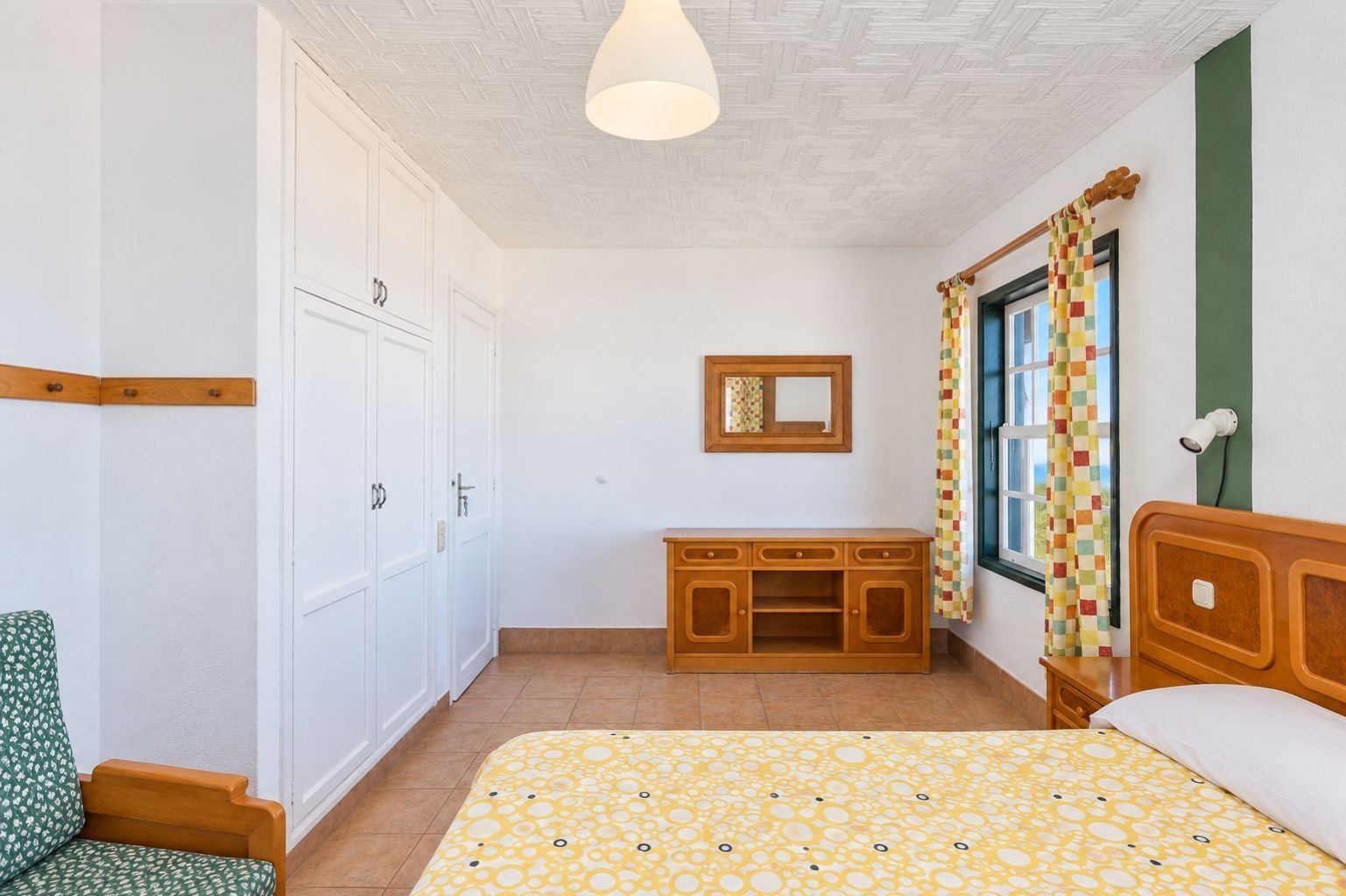 Een slaapkamer met een bed, een houten dressoir, een stoel, een spiegel en een raam met gordijn.