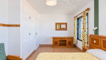 Een slaapkamer met een bed, een houten dressoir, een stoel, een spiegel en een raam met gordijn.