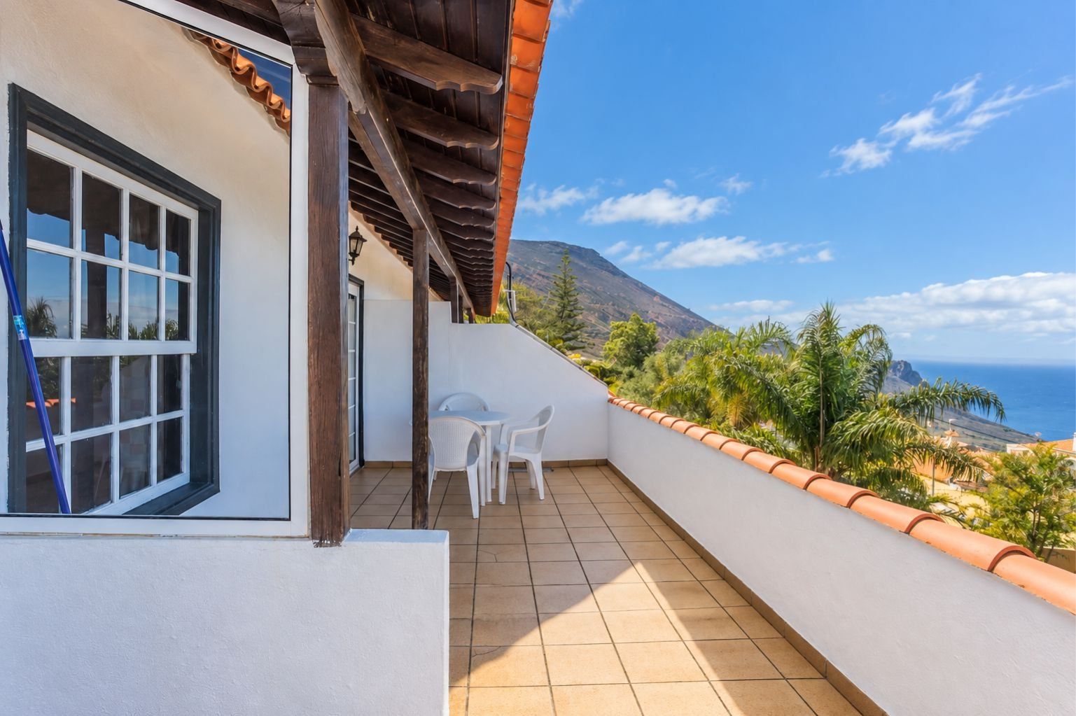 A balcony with a view of the sea and mountains, a white building, and a table with chairs.