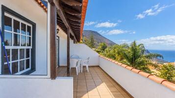 A balcony with a view of the sea and mountains, a white building, and a table with chairs.