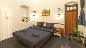 Habitación de hotel con una cama individual, una mesita de noche, un teléfono, una pequeña mesa, una silla, una puerta, una ventana y una lámpara de pared.